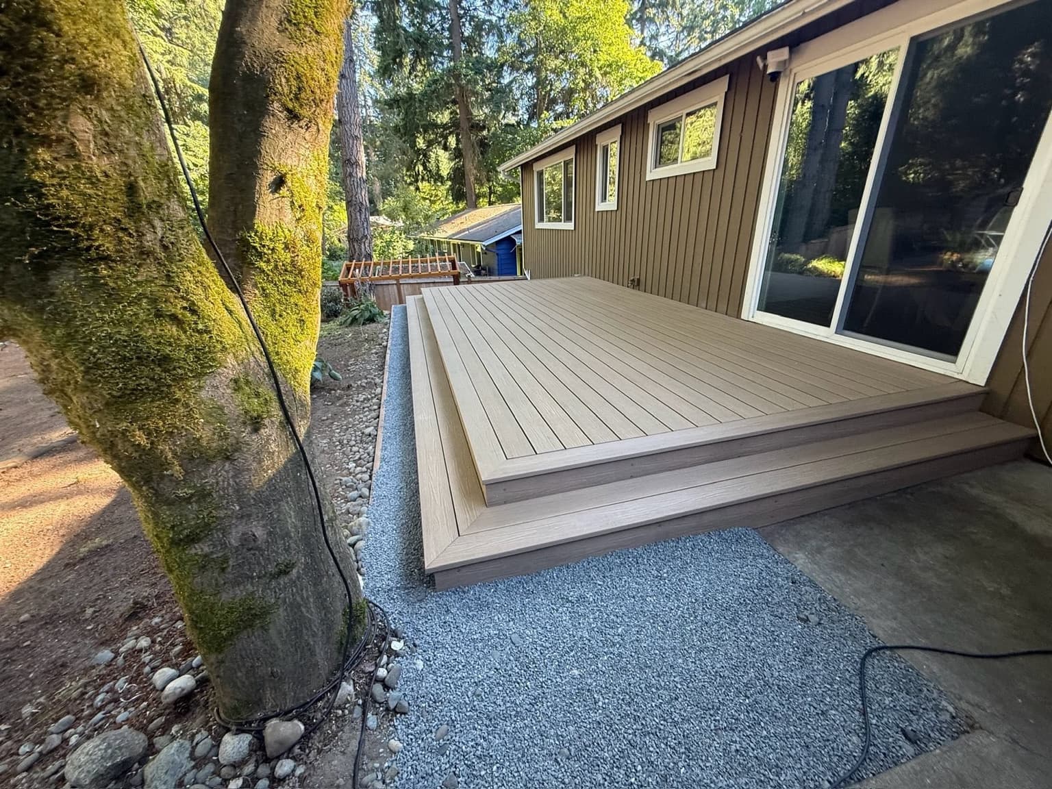 Low-profile tan composite deck with a wrapped full-width step and gravel drainage border, beside a mature tree