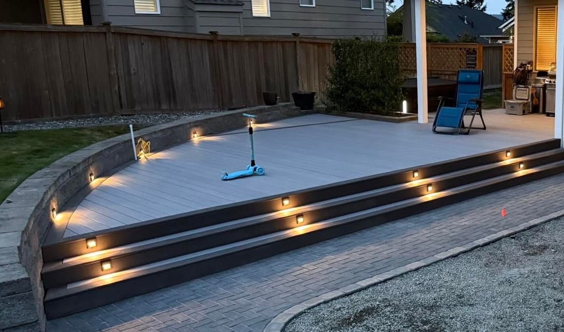 Curved composite deck with lit risers on every step, opening onto a herringbone paver patio at dusk, built by Best Pro Contractors